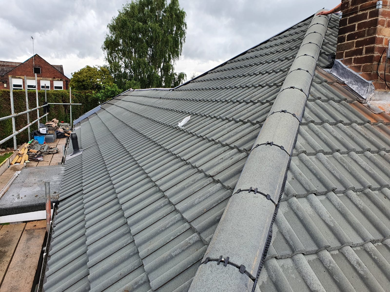 Re-bedded ridge line with mechanically fixed ridge clips running alongside a brick chimney stack on a tiled roof